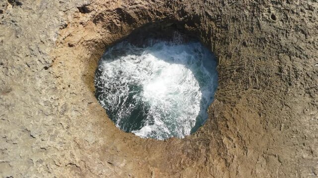Aerial parallax circles Watamula Hole in Curacao showing waves surging through natural rock blowhole with faint rainbow mist