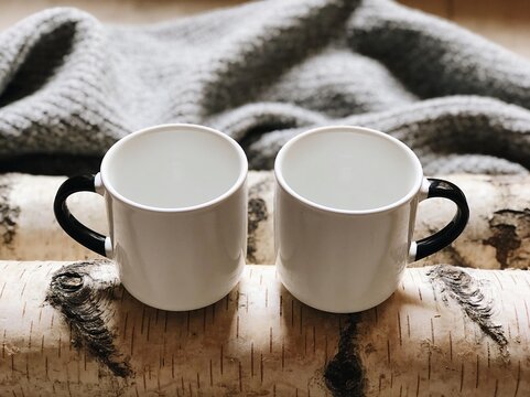 Mugs sit side by side on rustic birch wood logs draped with a soft grey knitted wool blanket.