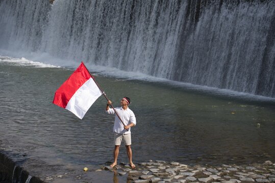 Man proudly holding the Indonesian flag at Tukad Unda Waterfall in Klungkung, Bali. Scenic riverside landscape celebrating patriotism with cascading water and a tropical natural backdrop.