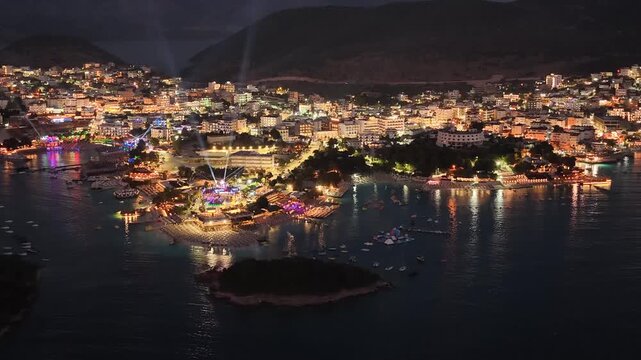 Spectacular night scenery of Ksamil, Albania captured by drone. The footage highlights the glowing city streets, lively beach resorts, and the dark silhouette of the famous Ksamil islands in the bay u