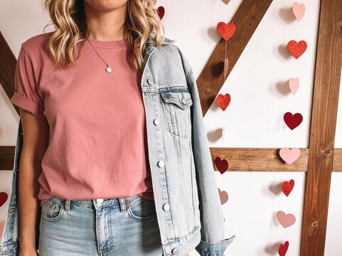Woman wearing a pink t-shirt and denim jacket stands before a decorative wall adorned with paper hearts.