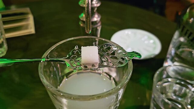 Close-up of absinthe louche service with sugar cube on a slotted spoon over a glass, water drop falling from fountain, cloudy drink, green bar lighting, and moody cocktail atmosphere.