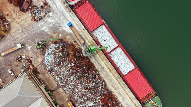 A large ship sits at the dock, unloading scrap metal into waiting containers. Nearby, cranes lift debris and materials from a pile on land. Workers follow safety procedures.