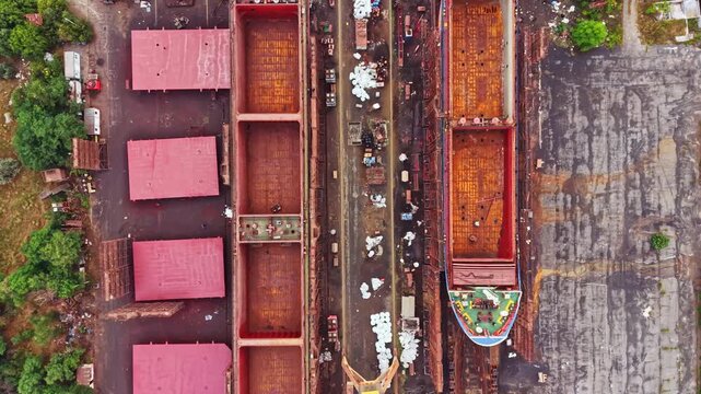 Workers are assembling a large ship in a shipbuilding yard. Surrounding buildings hold materials and equipment. Crane lifts parts as vehicles move around the site during a sunny day.