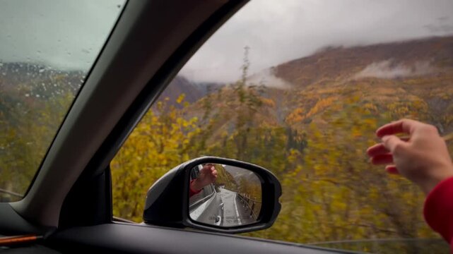 Traveler enjoys scenic drive through stunning fall foliage in mountains while waving hand outside vehicle. Adventure and exploration concept