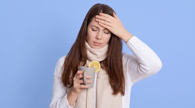 A young woman feeling unwell, holding her hand to her forehead and a mug with lemon, suggesting a cold or flu.