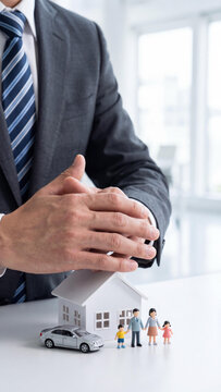 Close-up of a businessman in a suit protecting a white toy model house, a miniature silver car, and a tiny family figurine with his hands on a white desk in a bright office.
