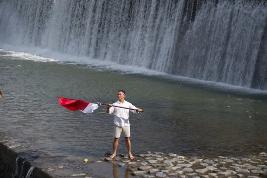 Man proudly holding the Indonesian flag at Tukad Unda Waterfall in Klungkung, Bali. Scenic riverside landscape celebrating patriotism with cascading water and a tropical natural backdrop.