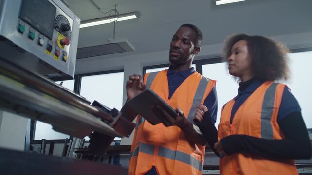 Two labour workers in industrial plant, looking at a tablet, discussing production and digital technology integration in factory operations.