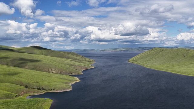 A breathtaking aerial drone flight over a peaceful mountain bay framed by rolling green hills and expansive freshwater. Towering clouds cast dynamic light across the landscape, creating a dramatic sum