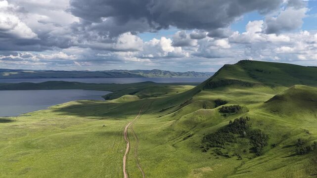 Cinematic drone footage soaring above rolling green hills under a dramatic cloudy sky. Soft sunlight and moving cloud shadows create striking patterns across the natural landscape, revealing the beaut
