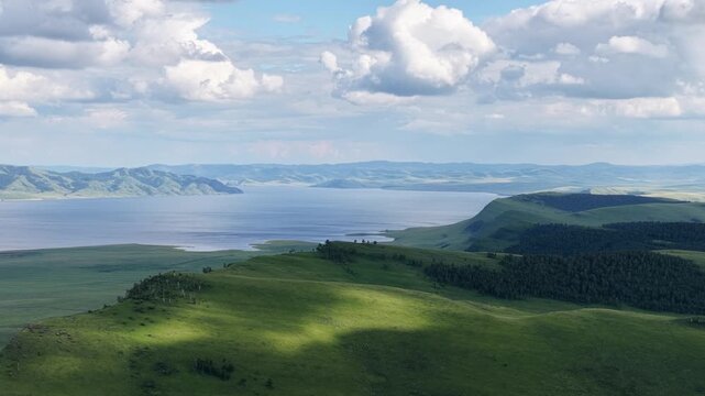 Spectacular drone footage capturing a vast mountain lake bordered by rolling green highlands and forested slopes beneath towering clouds. The changing light creates a peaceful yet dramatic atmosphere,