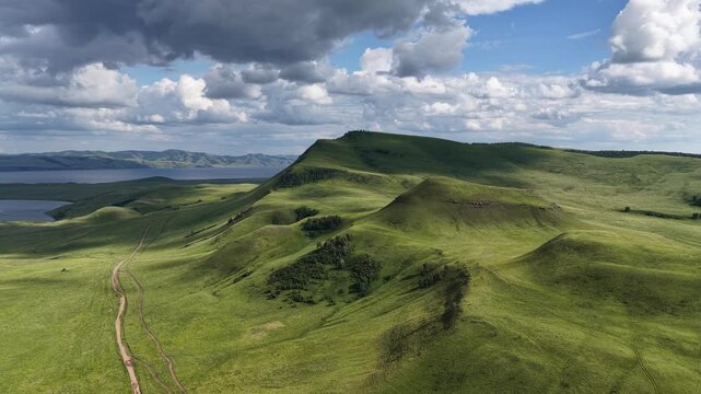 Cinematic drone footage soaring above rolling green hills under a dramatic cloudy sky. Soft sunlight and moving cloud shadows create striking patterns across the natural landscape, revealing the beaut