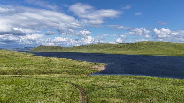 A breathtaking aerial drone flight over a peaceful mountain bay framed by rolling green hills and expansive freshwater. Towering clouds cast dynamic light across the landscape, creating a dramatic sum