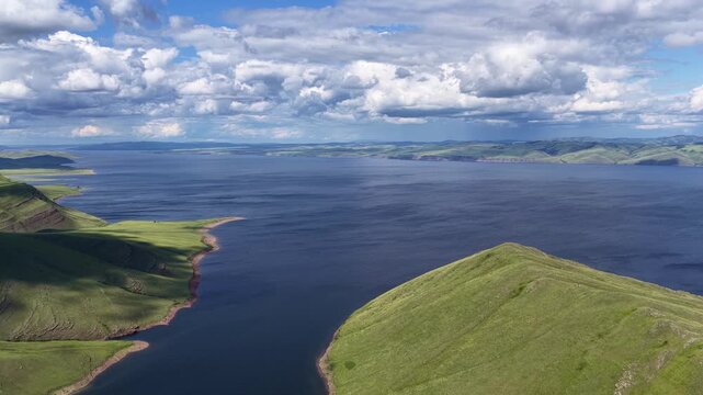 A breathtaking aerial drone flight over a peaceful mountain bay framed by rolling green hills and expansive freshwater. Towering clouds cast dynamic light across the landscape, creating a dramatic sum
