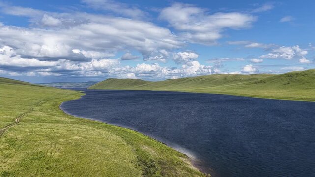 Cinematic drone footage of a tranquil mountain lake stretching between rolling green hills beneath dramatic cumulus clouds. Scenic aerial landscape featuring pristine nature, deep blue water, panorami