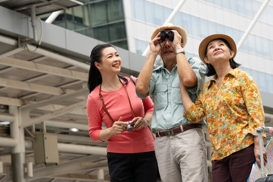 Happy Asian family group enjoying sightseeing city tour. Elderly parents with binoculars and map alongside their adult daughter with camera, looking up at modern architecture during an urban vacation.