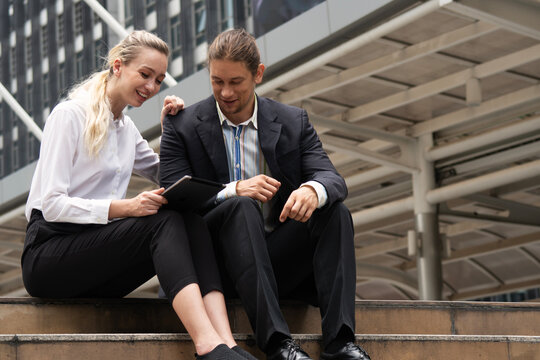 Smiling Caucasian business colleagues sitting on outdoor city stairs looking at a digital tablet. Happy professional team collaborating and discussing a project outside the office.