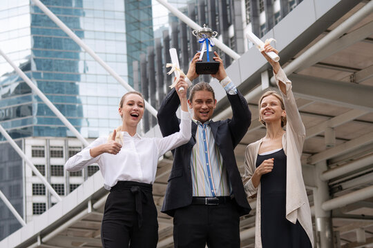 Happy Caucasian business team celebrating success in the city. Professional colleagues cheering while holding a championship trophy and award certificates for corporate victory.