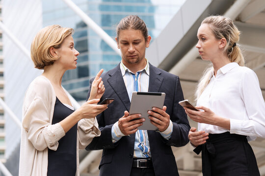 Serious Caucasian business team discussing a project outdoors in the city. Professional male manager holding a digital tablet while a female colleague points and explains a strategy.