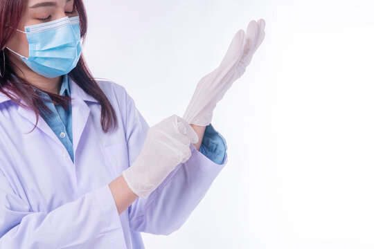 Asian female doctor or scientist wearing a medical face mask and white lab coat putting on sterile latex gloves. Healthcare professional preparing for clinical examination on a white background.