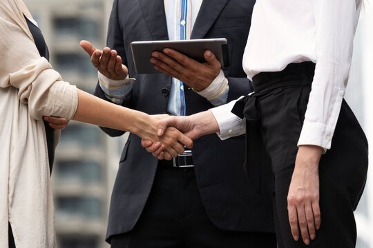 Close-up of professional business women shaking hands to seal a deal outdoors in the city. A male manager in a suit holds a digital tablet in the background. Corporate partnership and agreement.