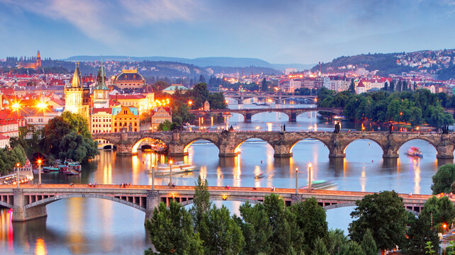 Twilight cityscape featuring multiple bridges over a river in Prague skyline, with historic buildings and street lights.