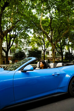 An Asian woman drives a blue convertible sports car down the street.