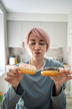 Thoughtful mature woman keeping healthy diet and weight, holding glazed doughnuts deciding on eating, pondering female coping with temptations to try junk food for meal, modern sugar addiction.