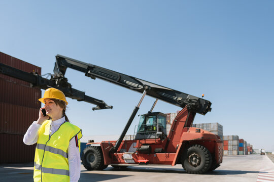 Female worker on the phone in front of crane with cargo container on industrial site
