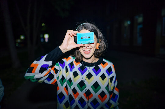Smiling music fan holding retro cassette over eyes walking at night