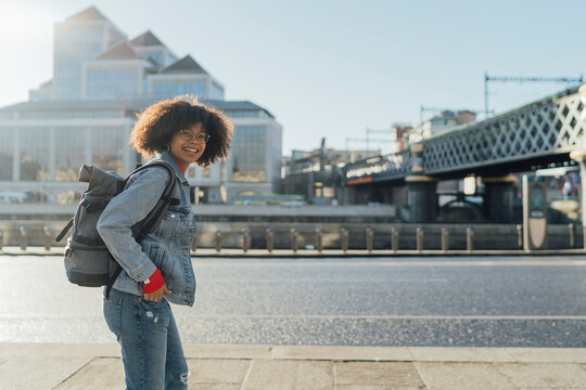 Smiling young woman with backpack standing on footpath by river in city