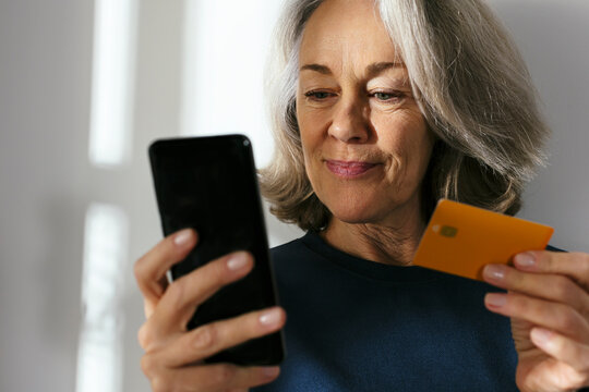 Mature woman with credit card using smart phone in front of wall