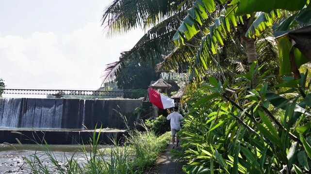 Man running while carrying the Indonesian flag near Tukad Unda Waterfall in Klungkung, Bali. Patriotic outdoor scene with tropical scenery, cascading water, and lush greenery.