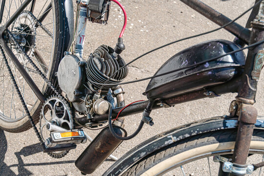 Motorized bicycle featuring two-stroke engine, mounted on rusted frame, showcasing mechanical details, connected to rear wheel via chain mechanism, set against concrete background