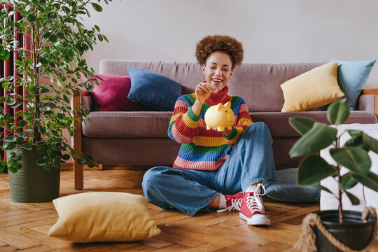 Happy young woman putting coin in piggy bank at home