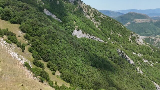 Pendii montani con vegetazione fitta e affioramenti rocciosi, vallate verdi e rilievi sovrapposti sotto un cielo azzurro. Paesaggio naturale ampio, luminoso e incontaminato