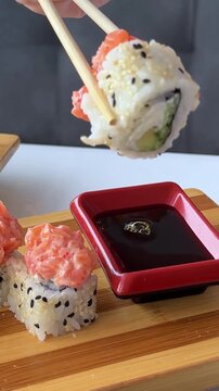 A specialty sushi roll covered with sliced salmon and black sesame is carefully dipped into soy sauce served in a red rectangular dish