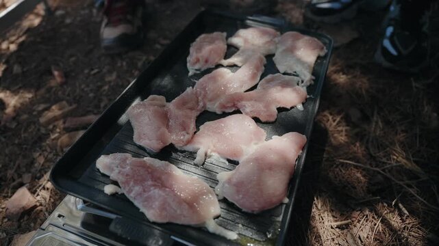 Fresh raw meat cutlets arranged on black tray over camp grill on dry ground outdoors