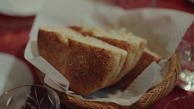 Sliced crusty bread in a napkin lined wicker basket on a dinner table with wine glasses