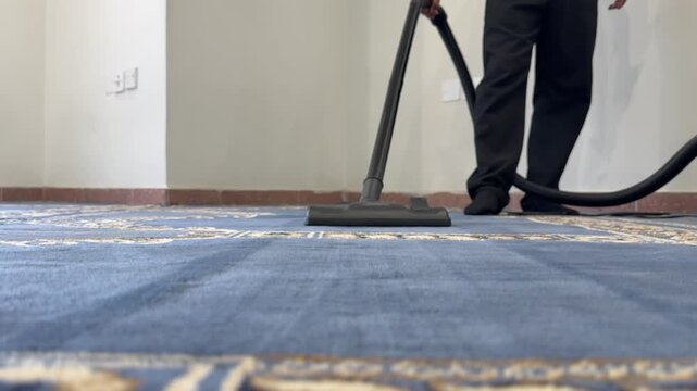 Low angle floor level shot of a man wearing black socks and pants vacuuming a blue patterned carpet. The worker cleans the floor inside a mosque or modern indoor space in Jabriya, Kuwait.