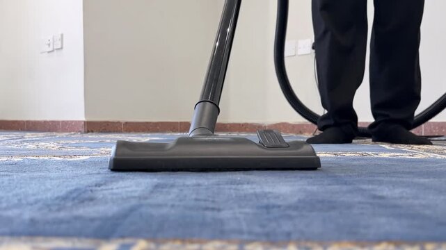 Close up floor level shot of a black vacuum cleaner head moving across a blue patterned mosque carpet. A janitor in black pants cleans the floor inside a bright room in Jabriya, Kuwait.