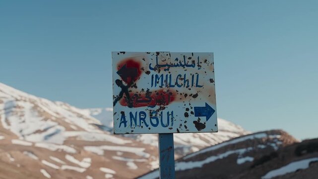 Rusty directional sign pointing to Imilchil and Anrgui with snow capped High Atlas peaks behind Morocco