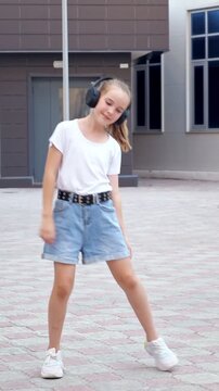 Cheerful young girl wearing large headphones listening to music and dancing happily on an urban street, enjoying her leisure time with energetic moves and a genuine smile