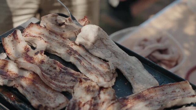 Hand turning sizzling pork belly strips on a hot black griddle with fork