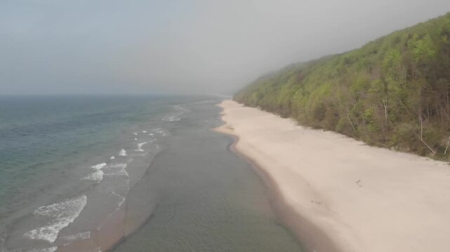 Wide drone footage tracks along a quiet sandy beach near Poddabie, with Baltic Sea surf on one side and green forested dunes fading into a hazy Polish coastline.