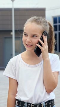 Cheerful smiling preteen girl with a ponytail using a smartphone for a phone call while standing outside. Modern childhood and communication technology concept for a young generation