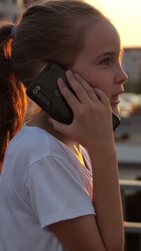 Young pretty girl talking on a smartphone outdoors during a beautiful golden hour sunset, showing a profile view of her face and highlighting her hair with a warm rim light