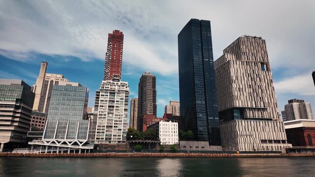 Boat glides past Roosevelt Island's East River waterfront featuring the Cornell Tech campus, residential towers and historic power station beneath dramatic clouds.