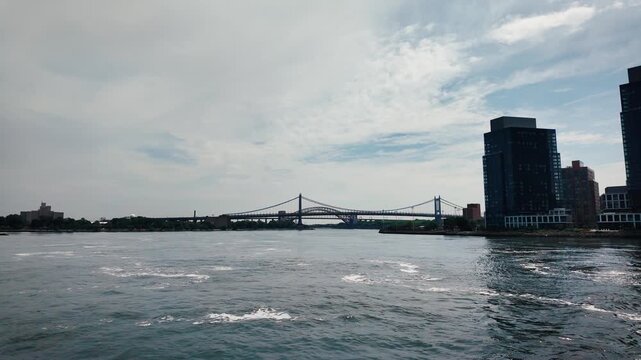 Cinematic boat approach toward the RFK Bridge with the Hell Gate Bridge behind, crossing New York's East River under dramatic clouds and calm water.